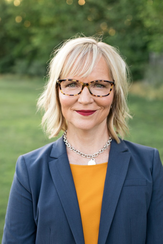 Upper-body Shot of Lisa Jones Christensan wearing a blue jacket and a yellow top. She stands in front of blurred outdoor background