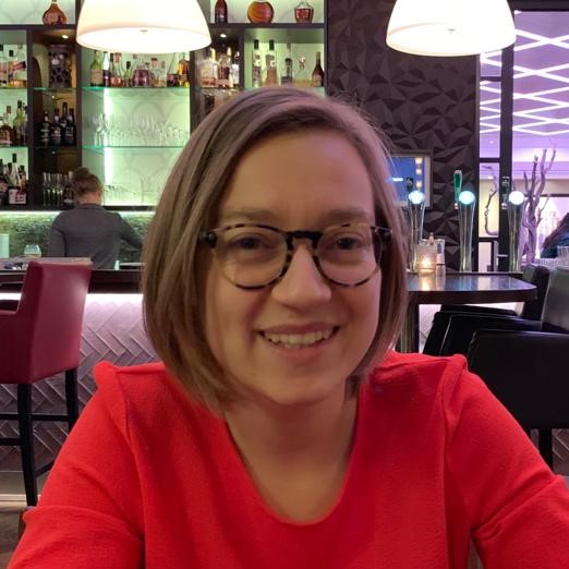 head shot of Ann-Sophie Baeken wearing a red top standing in front of a br with lots of bottles on glass shelves