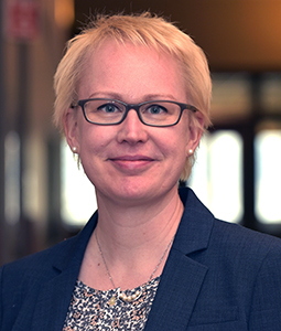Upper body shot of Maija Renko. Maija has blonde short hair and wears glasses. She stands in front of a blurred city background