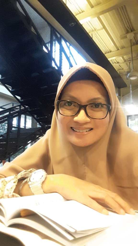 Upper body shot of Anis Wahyu Intan Maris wearing glasses and a headscarf. She sits at a desk in a large room. A book is on the desk and stairs are visible in the background
