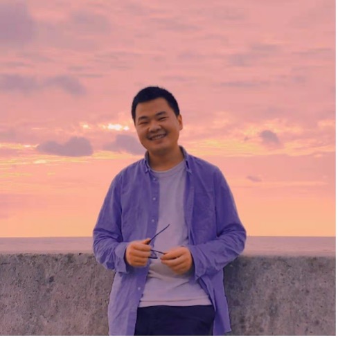 Upper body shot of Chi Zhang wearing a white T-Shirt and a blue open shirt over it. He is holding his glasses in his hands. THe background shows a landscape during sunset
