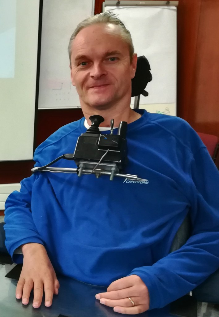 upper body shot of Vic McKinney wearing a blueshift. Vic is in a lecture room. His wheelchair and assistive technology is visible