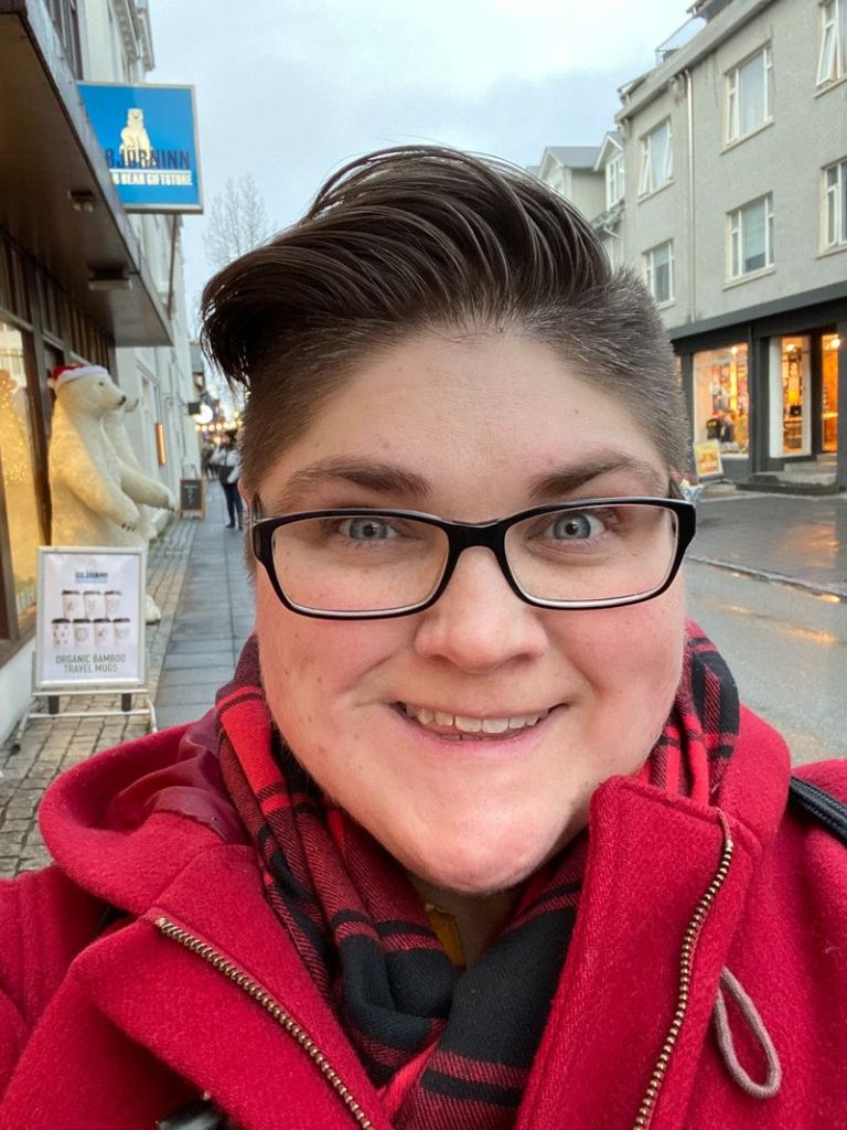 upper body shot of Kate Caldwell wearing a black top and a red jacket. Kate wears glasses. She is in front of a high street background