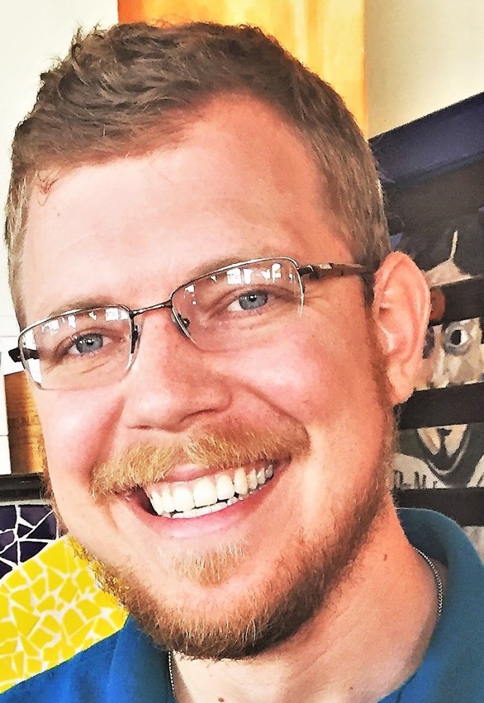 head shot of Thomas Dirth. He wears glasses and we can only see his head in the photo. He stands in front of a wall with different abstract posters