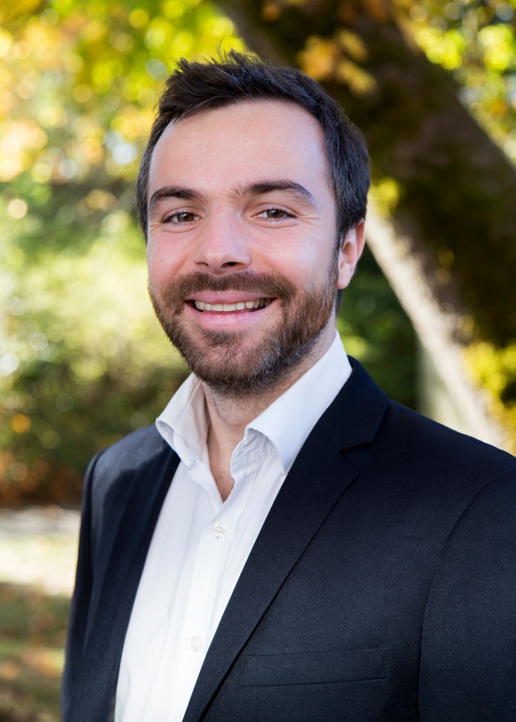Upper body shot of Camille Meyer wearing a white shirt and a black jacket. He stands in front of a blurred garden background