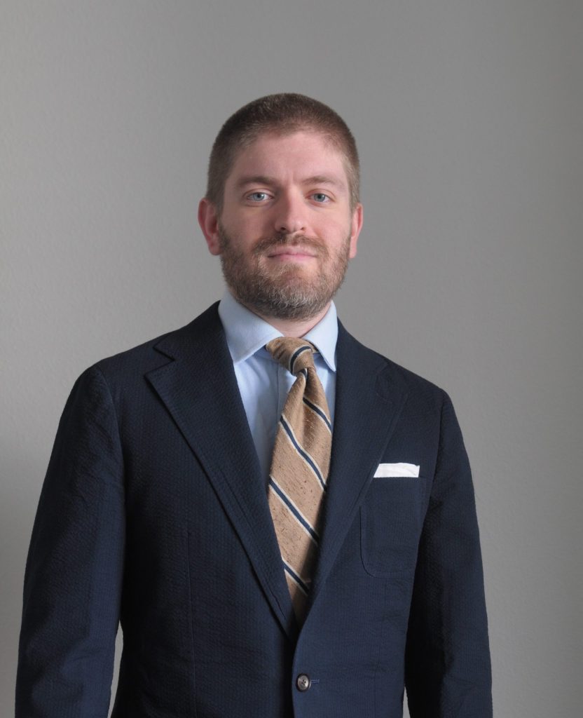 Uppoer body shot of Alexander C. Lewis wearing a shirt, tie and jacket standing in front of a grey background