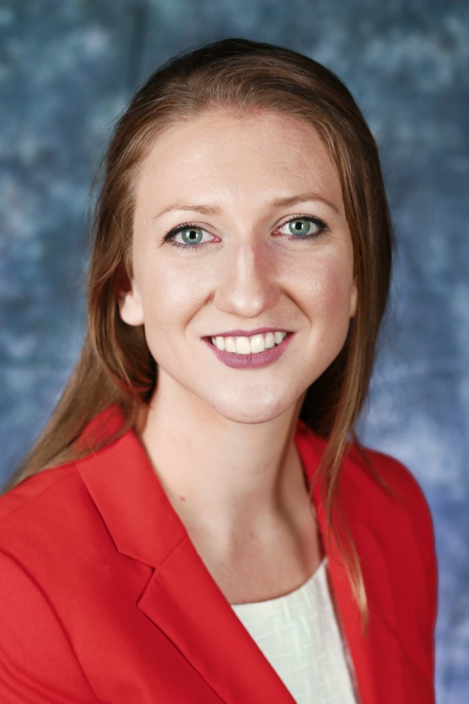 head shot of Christine Mihelcic wearing  a white top with a  red jacket. She is in front of a blue blurry background