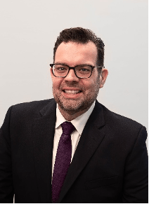 upper body shot of Christopher Sutton, a white man with short hair, wearing dark rimmed glasses and a suit. He has a big smile on his face