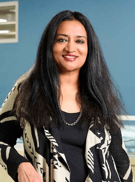 Head shot of Aparna Joshi , a woman with black long hair waring a black and white top in front of a blue background