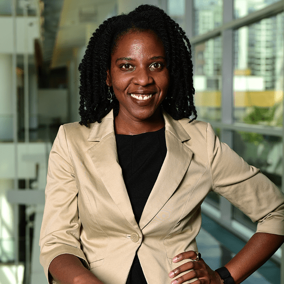 Upper body shot of Tiffany Johnson, a black woman with shoulder length hair wearing a white jacket and black top. She has a big smile on her face and stands in front of a blurred background of a building