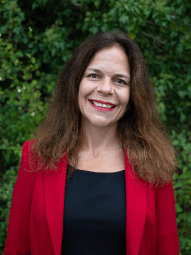 Upper body photo of Caroline Essers a white woman with dark brown shoulder long hair. She smiles at the camera and stands in front of greenery