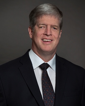 headshot of Kirk Adams a white man with grey hair wearing a black suit and tie in front of a black background