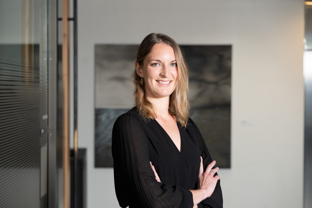 photo of Chloe Carmon , a white woman with dark blonde hair. She has her arms folded and stands at a slight angle to the camera. She wears a black top