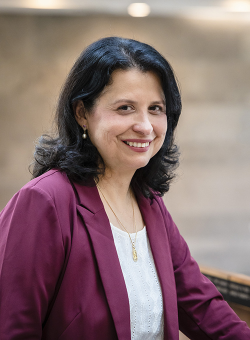 Upper body shot of Dusya Vera who has shoulder length dark hair. She is wearing a purple jacket and white top underneath