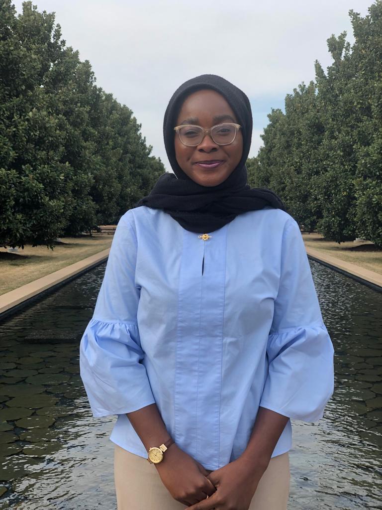photo of aminat-muibi, a black woman wearing a dark grey head scarf and a blue top. She stands in a park with a pond in the background.