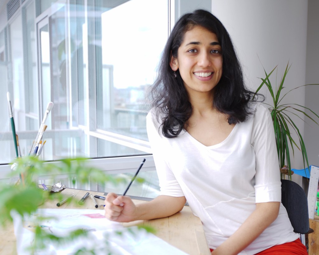 photo of Kanika Gupta who has long dark hair and wears a white top. She sits at a desk with arts materials