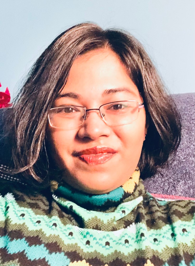headshot of Saheli Nath, a light skinned person with should long dark blonde, brown hair wearing glasses and a wooly jumper