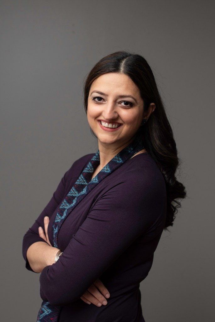 upper body tho of Maria Hamdan who has long black hair. She wears a dark jacket and a blue patterned top in front of a grey background. She smiles broadly