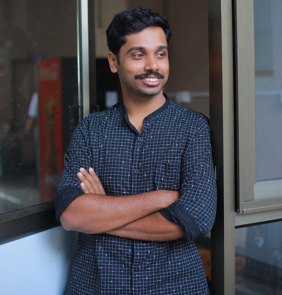 head shot of Riyas Kolopram Kandy, a man with brown skin and short dark hair and a mustachio. He wears a blue checkered shirt and has his arms folded. He smiles broadly slightly to the side.