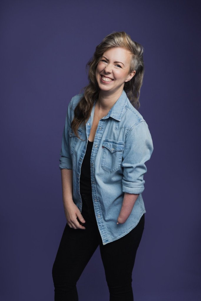 upper body shot of Alexis Hillyard, a woman with long brown hair falling over one of her shoulders. She wears a jeans jacket. It is clearly visible that her left arm has no hand. She wears black trousers