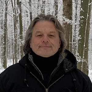 headshot of Ro’nikonkatste (Standing Strong Spirit) Bill Hill, an indigenous Canadian man wearing a lack coat. IN the background are snow covered trees. He has long dark and slightly grey hair