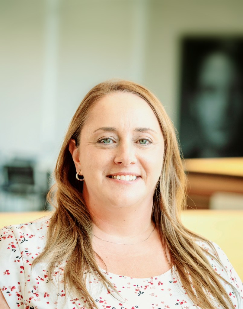 headshot of Sonja Bruschetto, a light skinned woman with long blonde hair. She ears a light top with a red pattern on it
