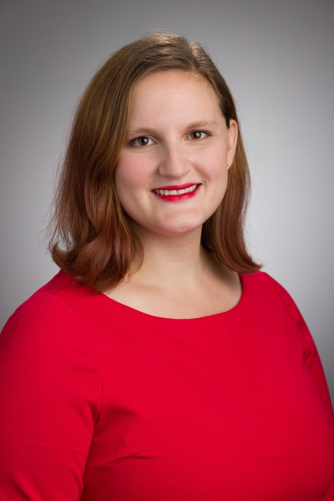 headshot of Samantha G Deane, a light skinned woman with long dark brown hair wearing a red top