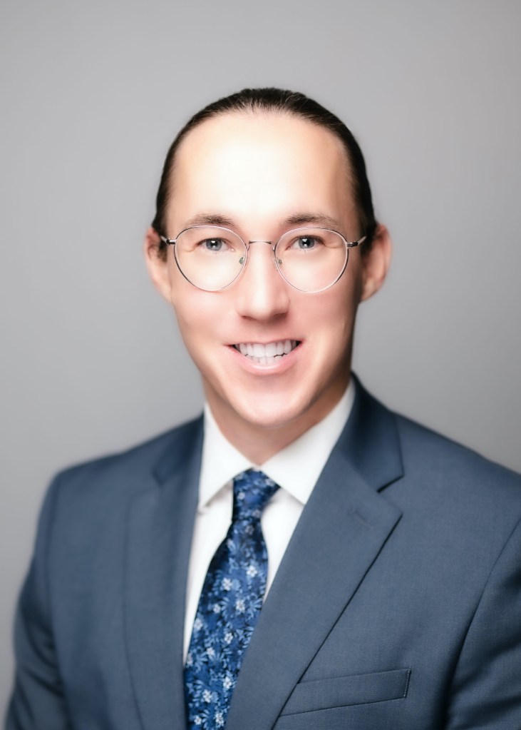 headshot of Walker Ray Dornisch, a light skin person with long dark hair tied back in a pony tail. Walker wears glasses