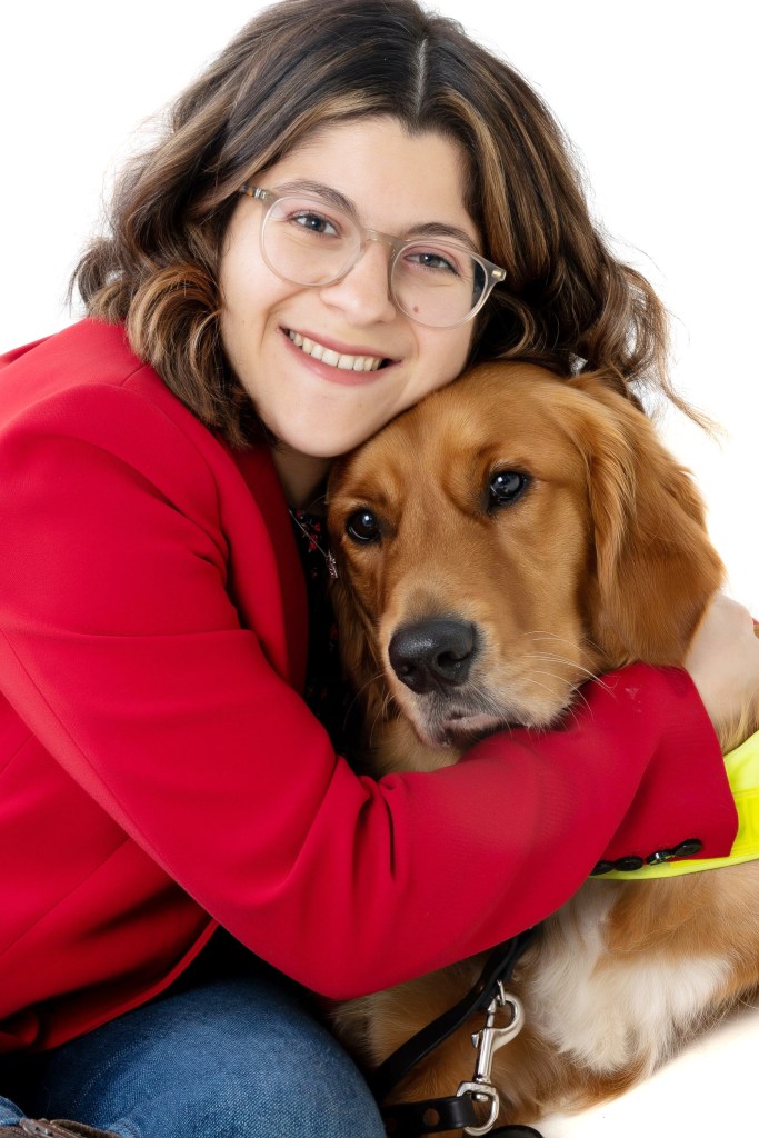 photo of Zein Al Maha Oweis wearing glasses and a red jumper. She cuddles her golden retriever guide dog