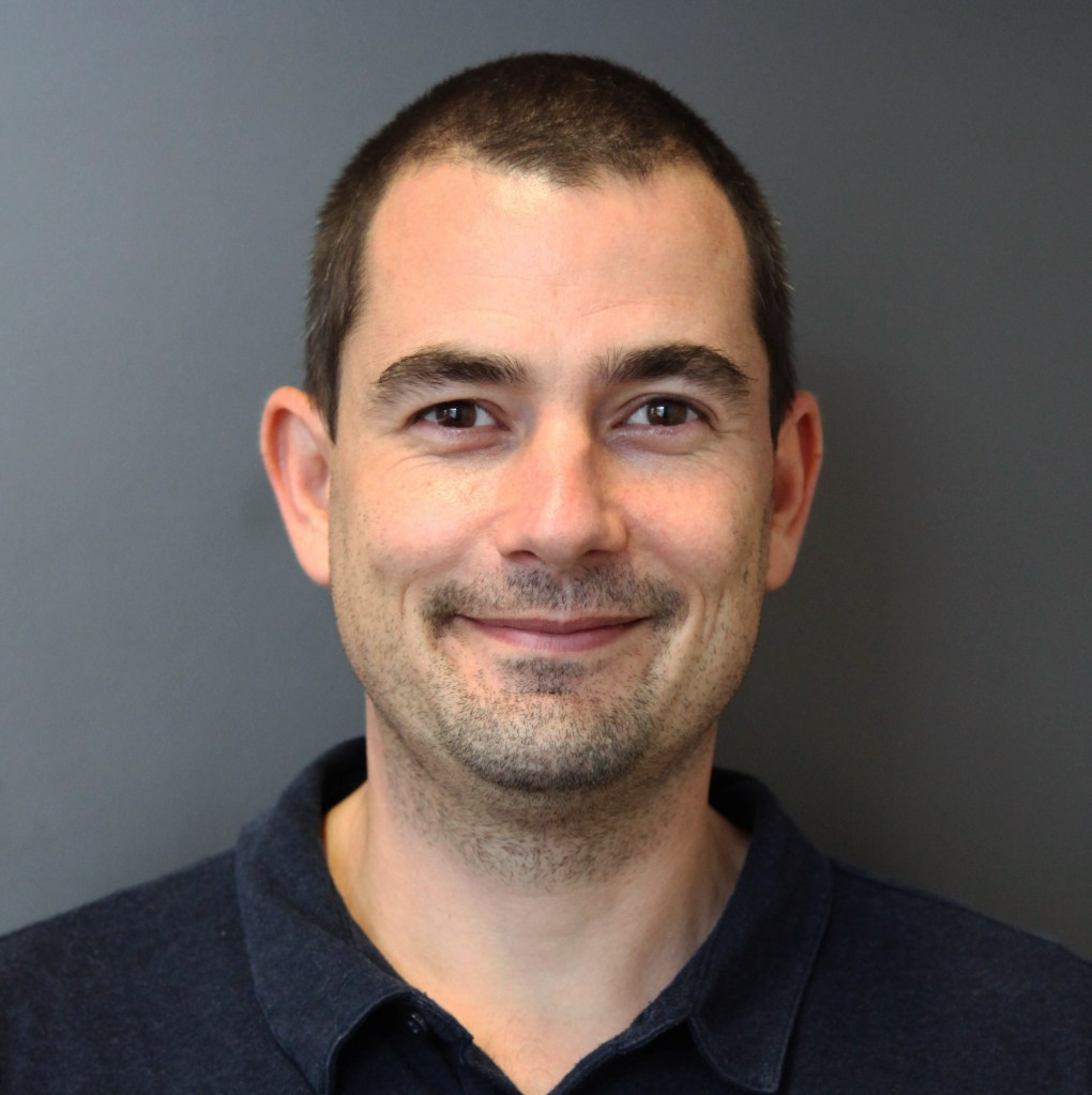 headshot of Johannes Krak, a person with olive skin, short black hair and a stubble. Johannes wears a black top