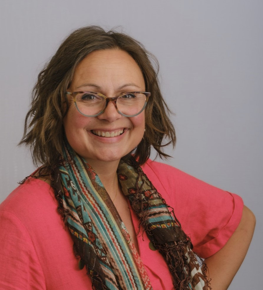 Headshot of Nancy Doyle, a middle-aged woman with black and brown hair wearing glasses. Nancy wears a pink top and has a colourful scarf wrapped around her