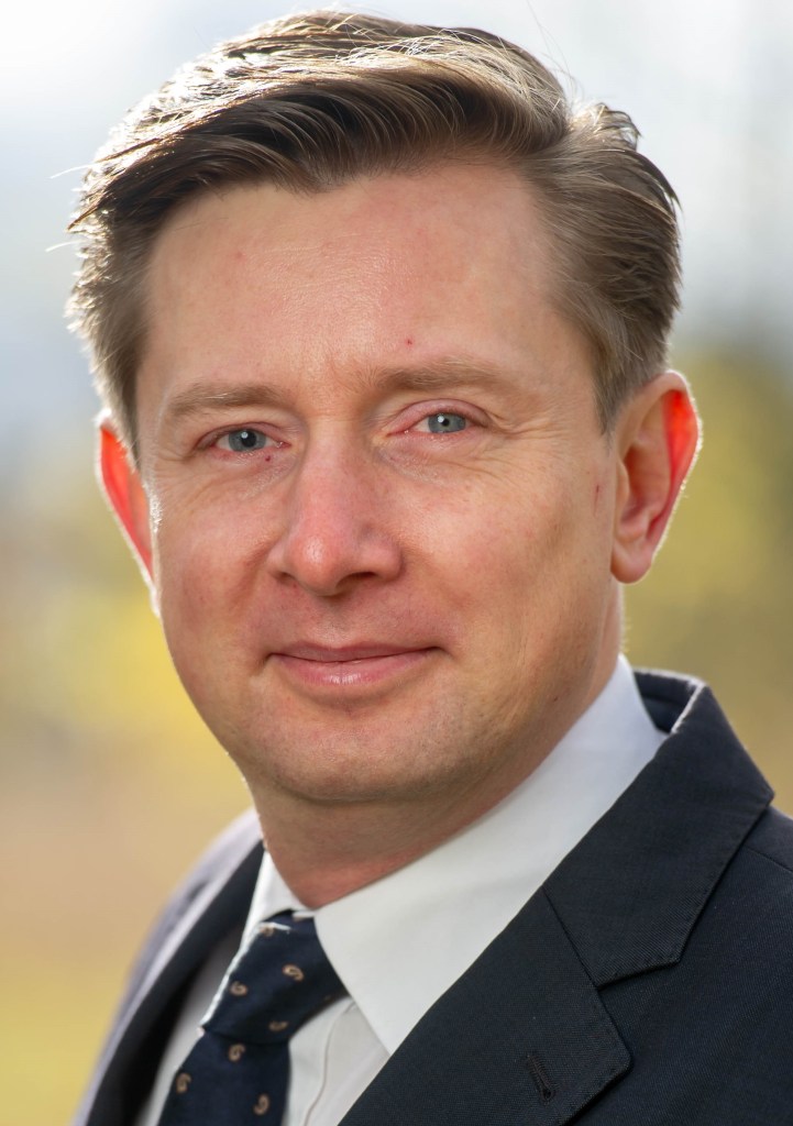 headshot of Felix Arndt a light skin man with short dark hair. He wars a suit and tie and smiles at the camera. The background is blurred. 