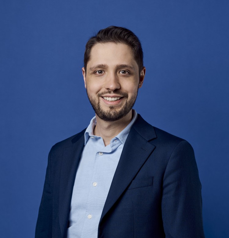 Headshot of Leandro Nardi, a white man with short brown hair. He wears a blue suite and a white shirt, standing in front of a blue wall. He smiles