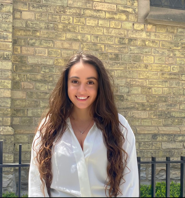 upper body shot of Rachel Pizzonia who has very long brown hair , wears a white shirt and stands in front of a yellow brick wall