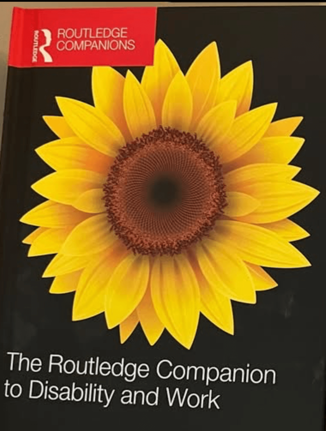 cover of the Routledge companion to disability and work. The cover is black with a yellow sunflower on it. the book title is in white writing. the Rutledge logo is in the top right corner with white on red writing
