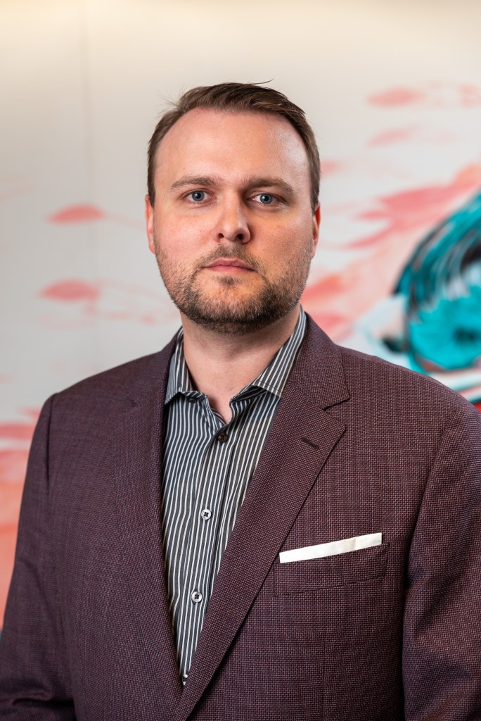 headshot of Andrew who has short dark hair and a beard. He wears a grey patterned shirt and a black jacket with a hancachief