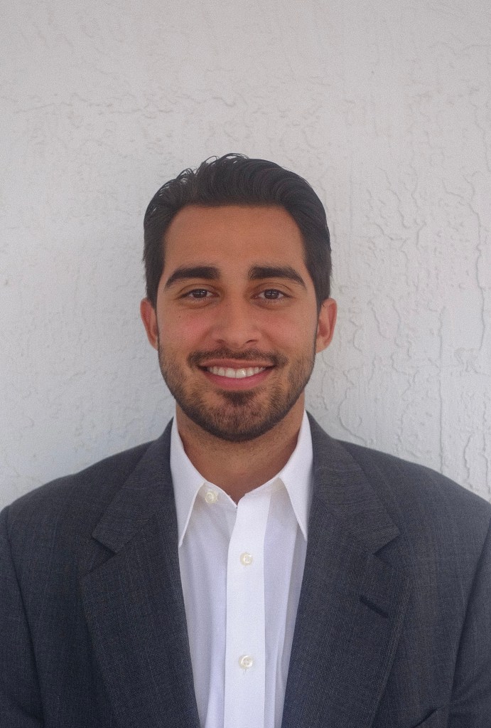 headshot of Julian. He smiles very broadly and wears a dress shirt with a black jacket over it. He has a short beard and moustache and short  ark hair.