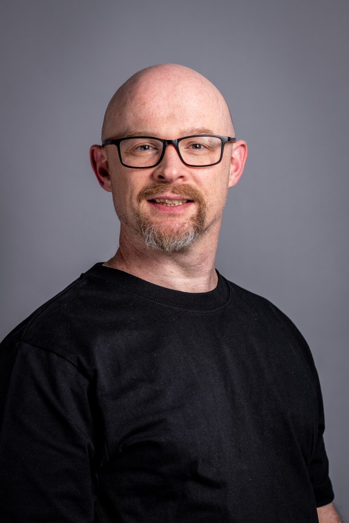 photo of Andy who is Bald white person with glasses, with a short dappled brown / grey beard, and wearing a black t-shirt. They are against a grey background.