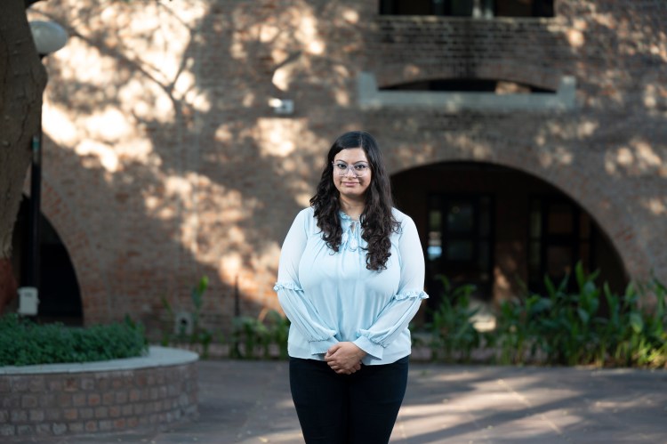 full body photo of Nikita who is a light skinned Indian woman with dark long hair who wears a light coloured long sleeve top with black trousers and stands in front of a big tree