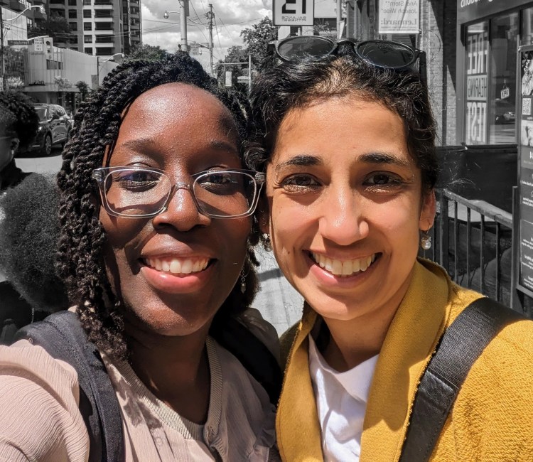 head shot of two women, one black and one brown, smiling brightly at the camera . they seem to be on a shopping street or in a city centre