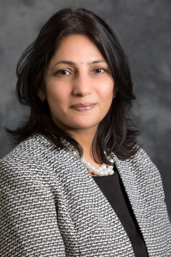 headshot of Tech who has shoulder long dark hair. She wears a grey patterned jacket and a black top underneath