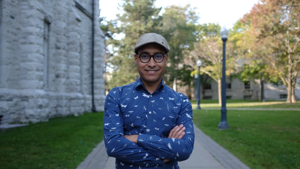 Photo of Alan Santinele Martino, a black man wearing a blue shirt with patterns on it. He also wears glasses and a cap