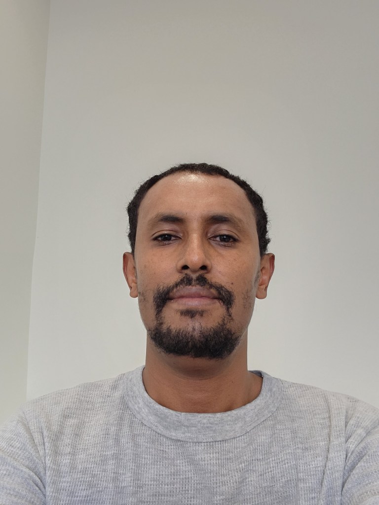 Photo of Dessalagn a black man with short hair and a bear wearing a grey top. 