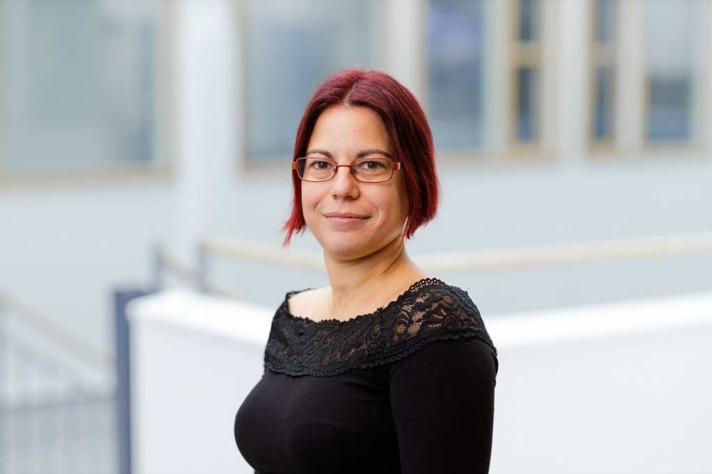 eadshot of Vasiliki Mylonopoulou. She is a white woman with dark red short straight hair. She wears a black t-shirt and glasses. The background is white and blurred.