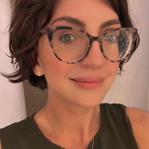 Headshot of Ren Lovegood. They are a white, woman presenting person with dark brown hair that goes to their chin. They are wearing large glasses and gold earings, The background is muted. 