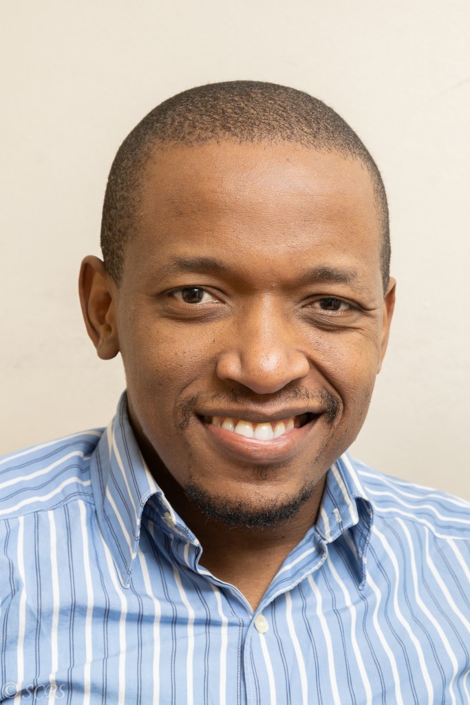 Headshot of Bongani Mapumulo. He is a black man with closely cropped hair and light facial hair. He is smiling and wearing a light blue striped button-down shirt. The background is plain and neutral.