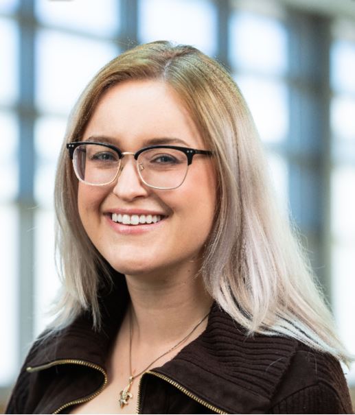 Headshot of Kayla N. Kozak. She is a white woman with long blonde hair and glasses. She wears a brown jacket and a necklace with a fleur-de-lis symbol. The background is a set of blurred windows. 