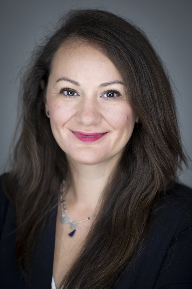 Headshot of Neva Bojovic. She is a light-skinned woman with long dark brown hair. She is smiling, wearing pink lipstick, a dark jacket, and a layered silver necklace. The background is plain gray.