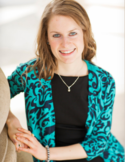 Headshot of Maura Danehey. She is a white woman with brown-blonde hair wavy hair, blue eyes, freckles, and a smile. She is wearing a bright blue sweater with dark-blue flowers, a silver necklace over a black dress. She is leaning on a pillar, in front of a tan background.