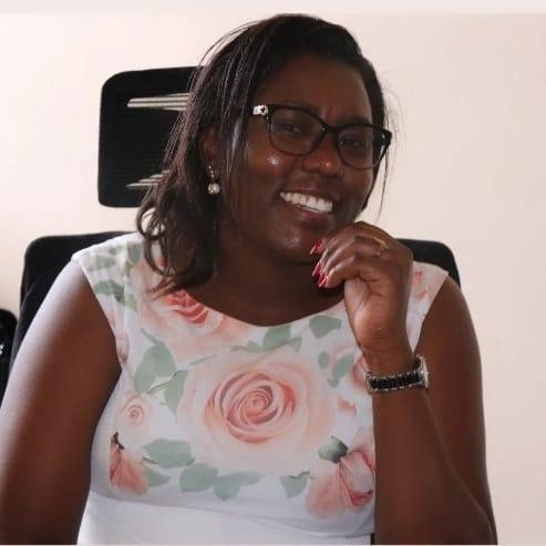 Headshot of Nazarene Makena, a Black woman, smiling while seated in an office chair. She has shoulder-length dark hair and wears glasses and pearl earrings. She is dressed in a white sleeveless dress with a soft pink and green floral pattern and a dark wristwatch on her left wrist. Her hand rests near her chin, and the background is plain and uncluttered.
